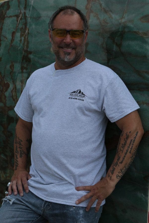 Middle-aged man with tattoos on his arms, wearing sunglasses and a white t-shirt, standing confidently in front of a quartz countertop backdrop.