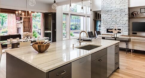 Modern kitchen interior with stainless steel appliances, an island from a renowned countertop company, and pendant lights, opening into a living area with a fireplace.