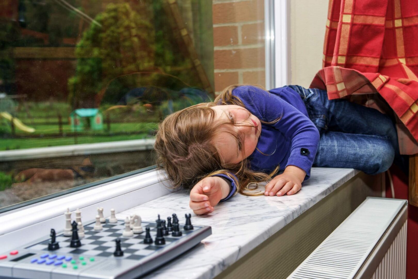 A young child asleep on a windowsill next to a chessboard, with remnants of a red blanket partially visible on the right.