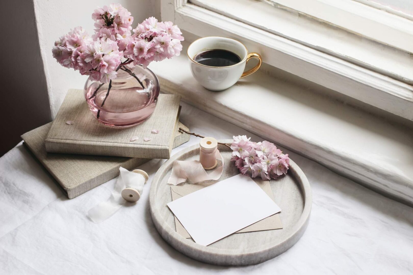 A cozy setting by a window with a vase of pink blossoms, a cup of coffee, books, and stationery on a tray atop countertop remnants.