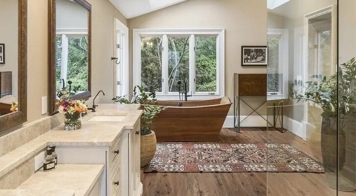 Modern bathroom with wooden freestanding tub, two vanities from a premier countertop company, rug, and large window with a garden view.