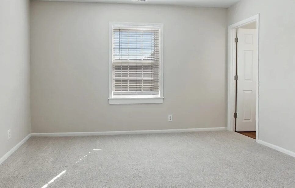 Empty room with beige carpet, a single window with blinds, and an open door on the right side.