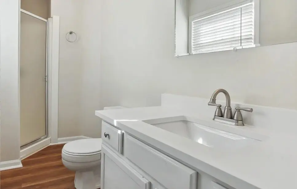 A clean, modern bathroom with a white vanity, sink, and toilet, next to a shower stall visible in the background.