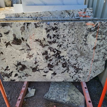 A countertop slab of granite with a mottled brown and white pattern, marked with red lines, resting on metal supports outdoors.