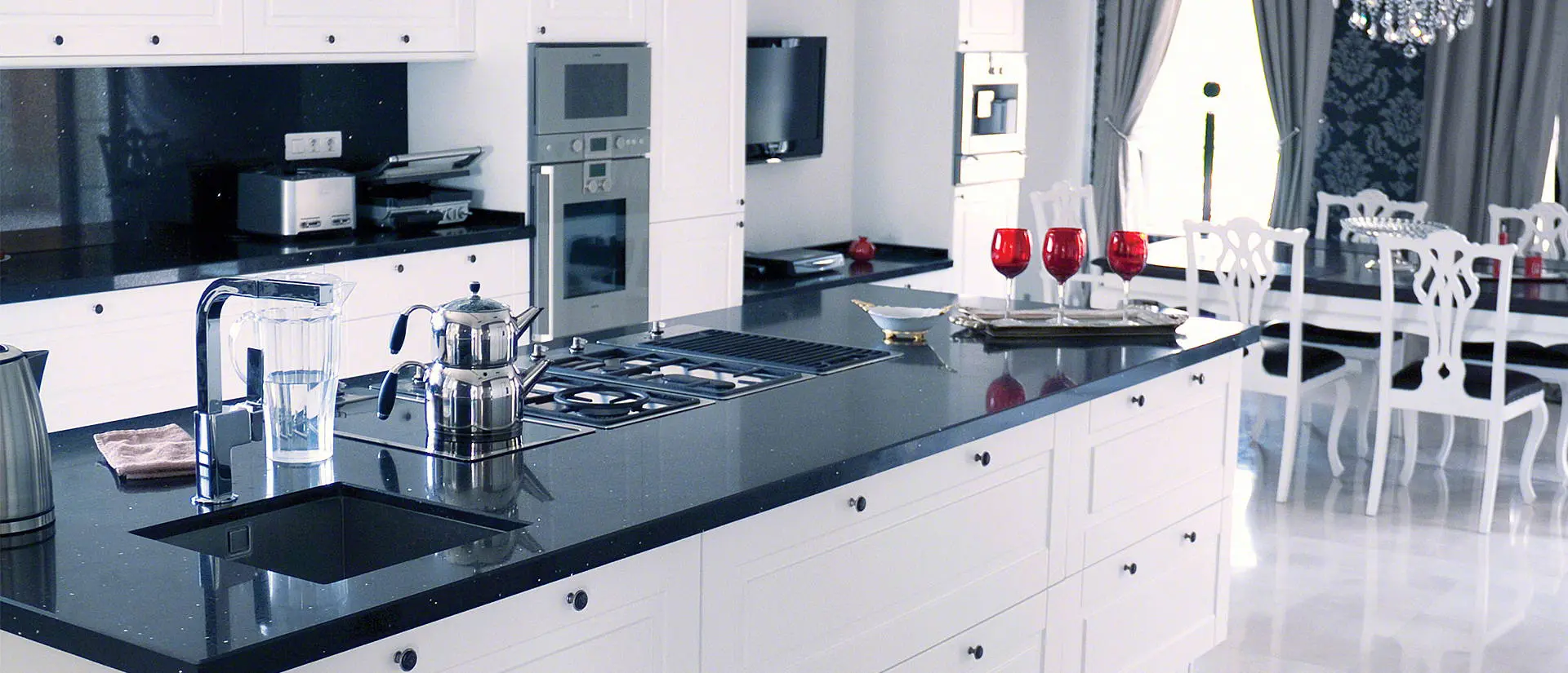 Modern kitchen with natural stone countertops, white cabinets, and stainless steel appliances. A dining area with white chairs and a table is visible in the background.